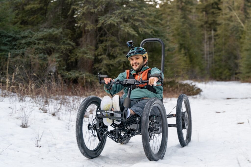 Man with limited hand function riding a four wheeled adaptive mountain bike in the snow. He is wearing winter gear and smiling.