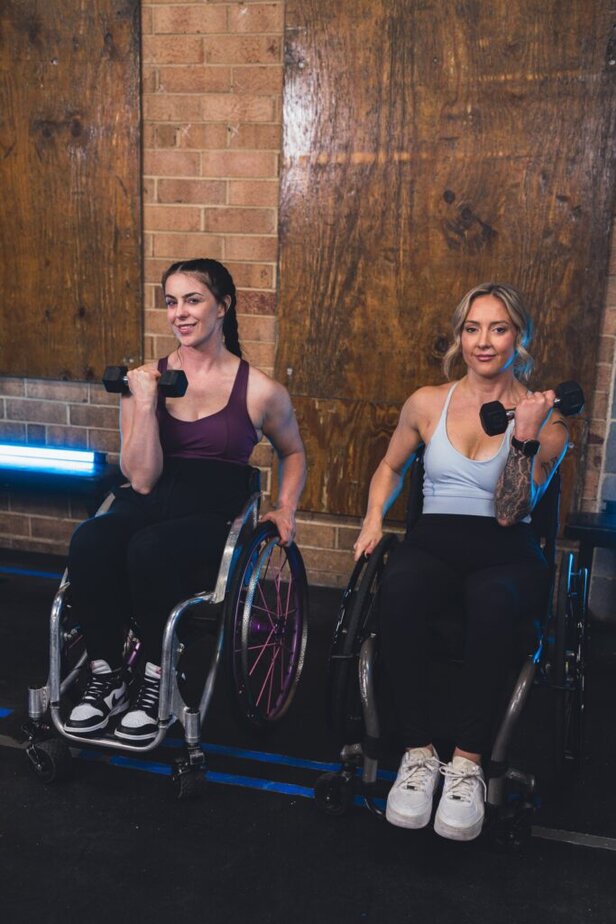 two female wheelchair users in gym, holding dumbbells in one hand.