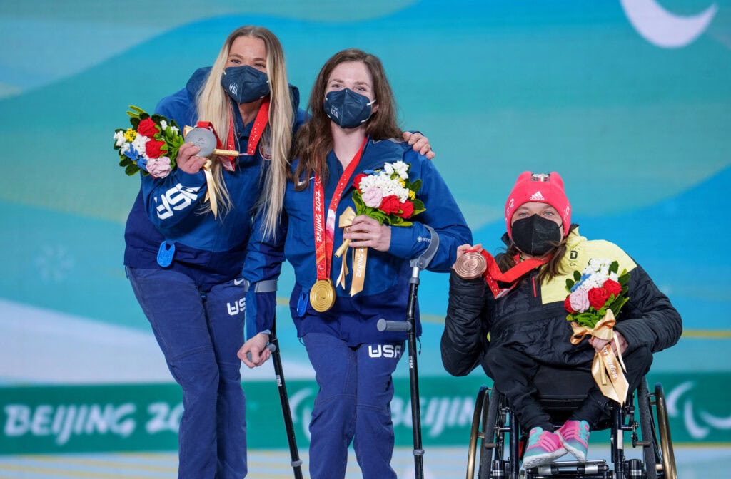 three woman, two using crutches and one using a wheelchair, pose with paralympic medals around their necks. 
