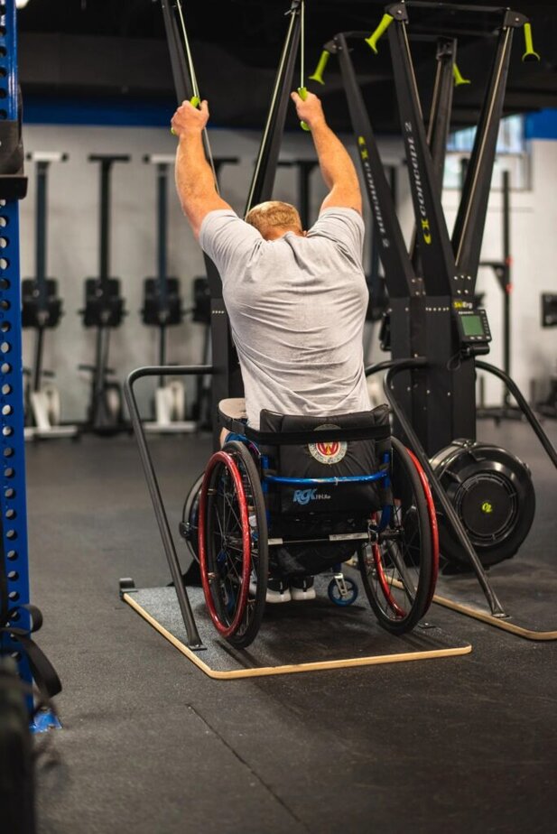 male wheelchair user with arms overhead pulling on cables from a SkiErg machine in a gym.