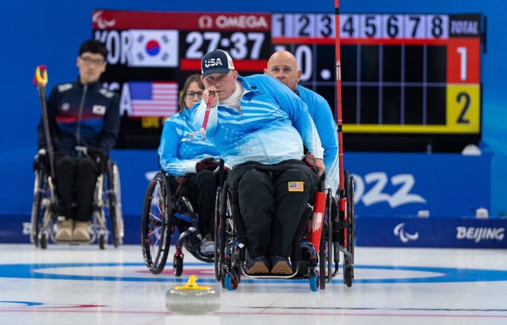 male wheelchair user on ice, watching a curling stone he just pushed.