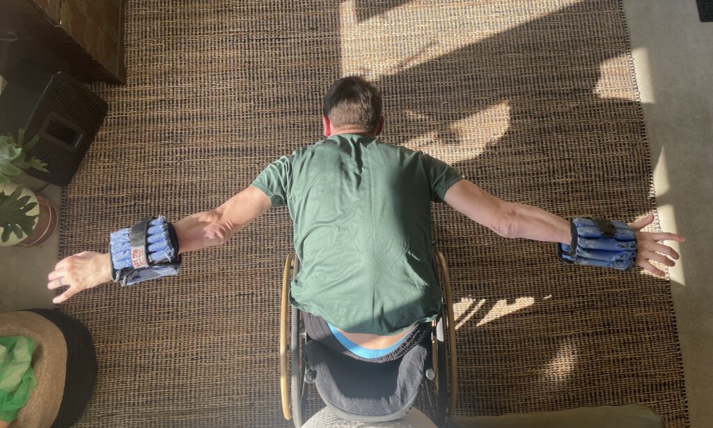 male wheelchair user leaned over his lap,, doing a back fly with wrist weights on his arms, photo is from overhead.