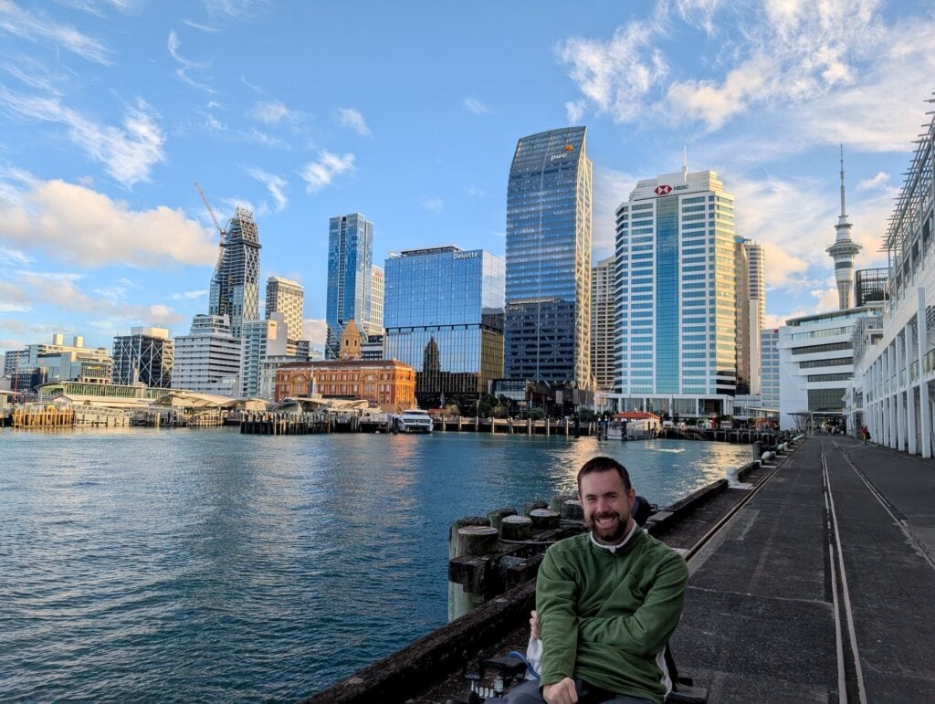 White man using power wheelchair sitting at a harbor with skyscrapers behind him.