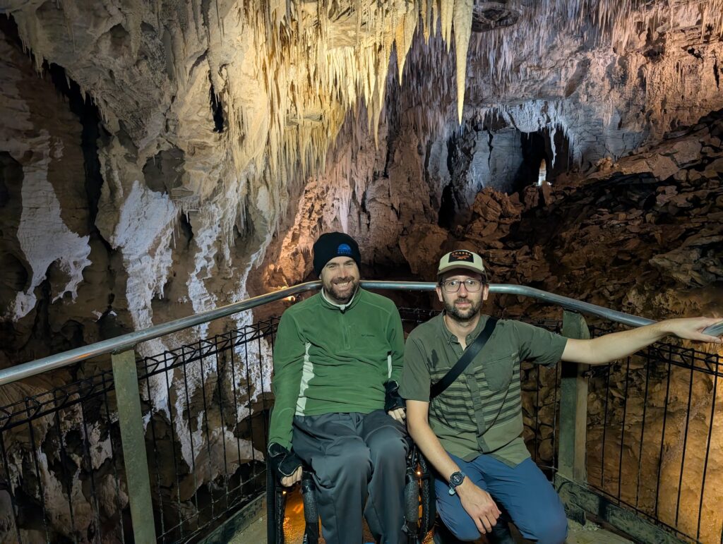 two men, one using a power wheelchair and one kneeling, pose in a cavern.
