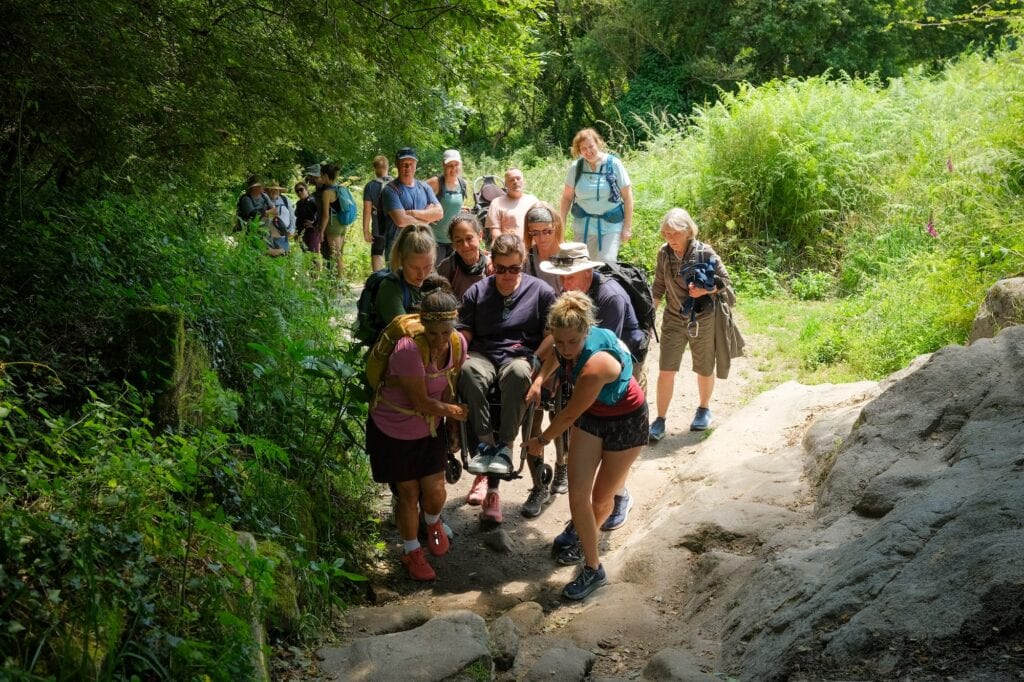 woman using a manual wheelchair being carried by a large group of people up a rocky trail.