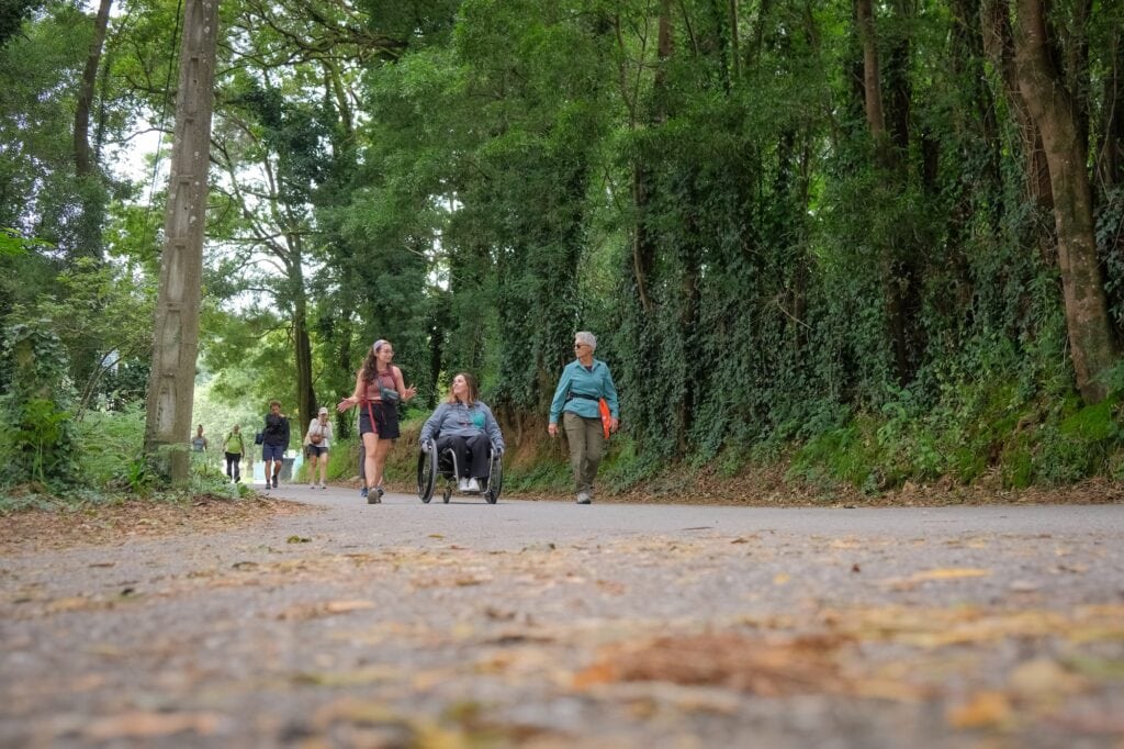 three women, one using a manual wheelchair, strolling on a paved forest path.