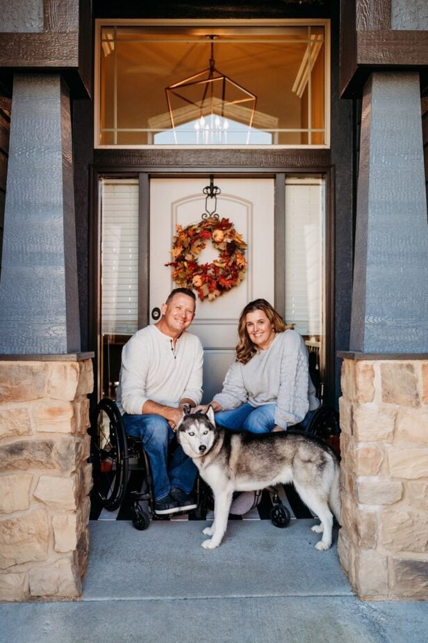 man and woman, both manual wheelchair users, pose with a husky in front of their house door. 