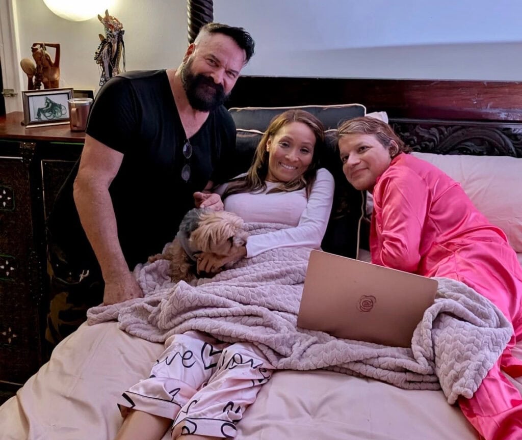 woman lying in bed with small dog on lap and a man on either side of her, all smiling at camera