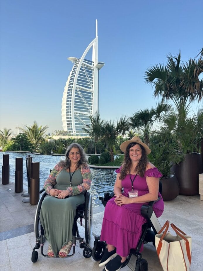 two women in wheelchairs sitting in front of large pool of water in Dubai with hotel in background