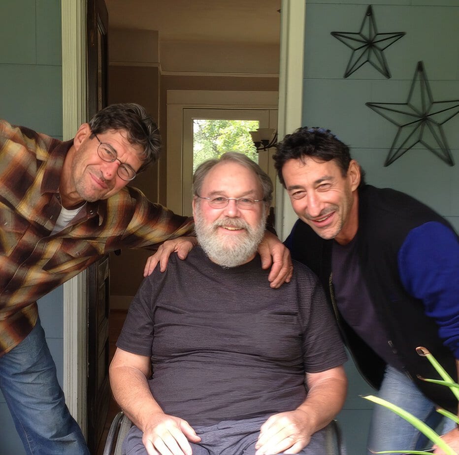 Man in wheelchair in with men standing on either side, hands on his shoulders