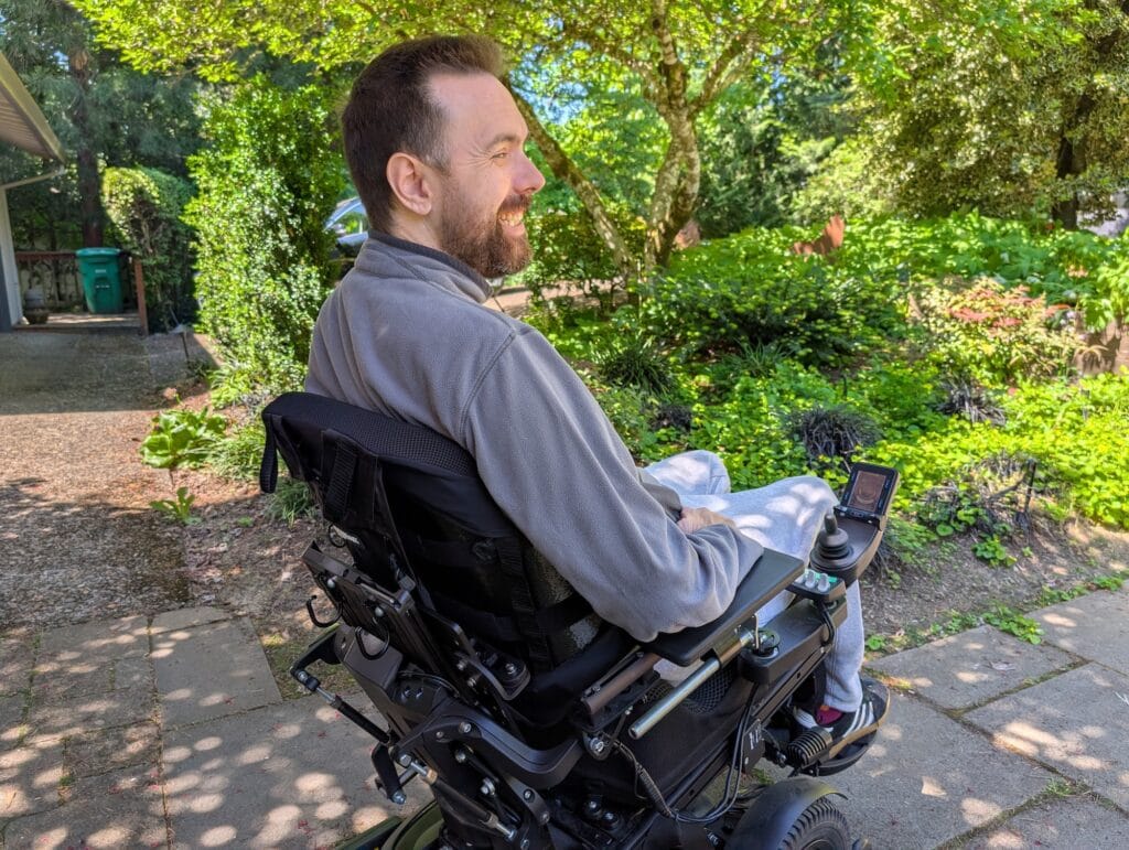 A man using a power wheelchair viewed from the side and slightly behind, showing a Bondar backrest attached to his chair. He is outside and smiling