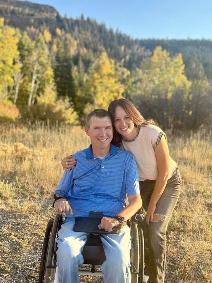man using a wheelchair and a woman putting her arm around his shoulder. The are in a field with trees behind them.