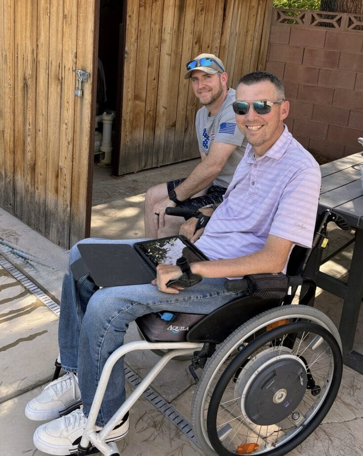 man in wheelchair on a patio with tablet on lab and man sitting behind him at a picnic table