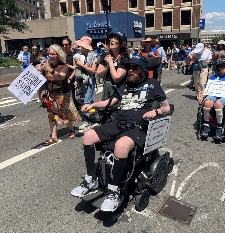 Woman walking in street next to man in powerchair at a disability rights demonstration