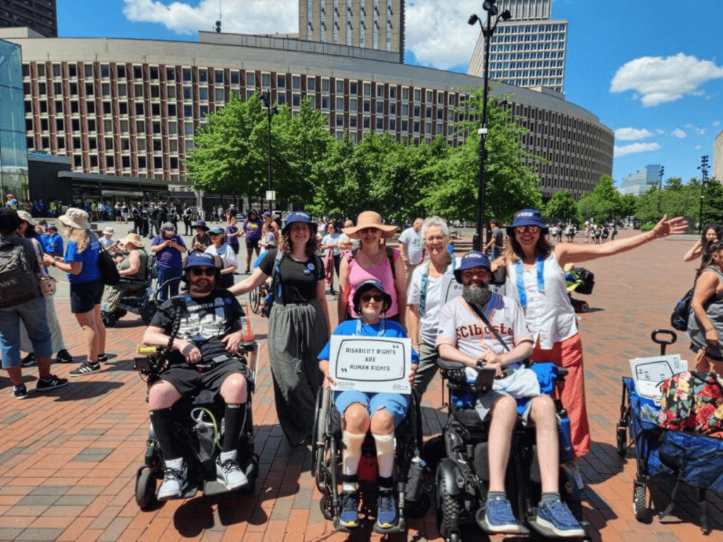 three wheelchair users with a group of people at a disability rights demonstration